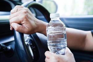 asiatische fahrerin hält kaltes wasser zum trinken im auto, gefährlich und riskiert einen unfall. foto