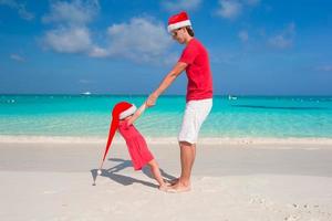 Kleines Mädchen und glücklicher Vater in Weihnachtsmütze haben Spaß am tropischen Strand foto