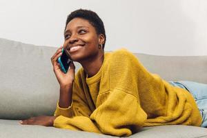 afrikanische frau, die zu hause auf dem sofa sitzt und handy benutzt foto