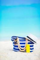 Strandzubehör - blaue Streifentasche, Strohhut, Sonnenbrille am weißen Strand foto
