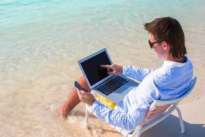 junger Geschäftsmann, der Laptop und Telefon am tropischen Strand benutzt foto