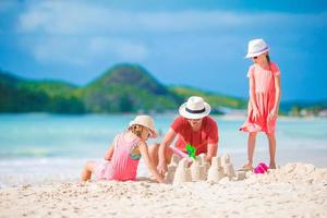 Familie macht Sandburg am tropischen weißen Strand. Vater und zwei Mädchen spielen mit Sand am tropischen Strand foto