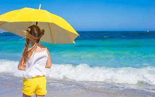 kleines Mädchen mit großem gelbem Regenschirm, das am tropischen Strand spazieren geht foto