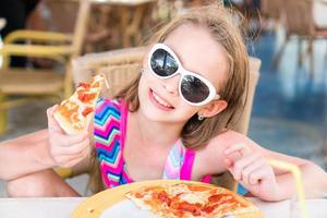 Porträt eines süßen kleinen Mädchens, das am Esstisch sitzt und Pizza isst foto