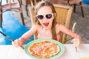 Porträt eines süßen kleinen Mädchens, das am Esstisch sitzt und Pizza isst foto