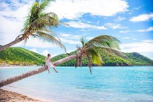entzückendes kleines mädchen, das während der sommerferien auf der palme am weißen strand sitzt foto