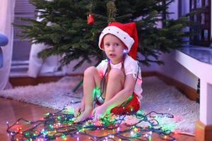 entzückendes kleines mädchen in weihnachtsmütze, das unter dem neujahrsbaum zwischen girlanden sitzt foto