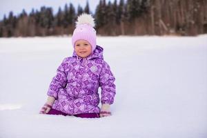 Kleines entzückendes Mädchen, das am sonnigen Wintertag auf dem Schnee sitzt foto