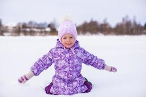 Kleines entzückendes Mädchen, das am sonnigen Wintertag auf dem Schnee sitzt foto