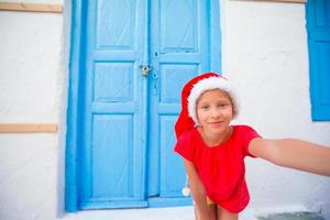 entzückendes kleines mädchen in weihnachtsmütze, das selfie in der schmalen straße im griechischen dorf macht foto