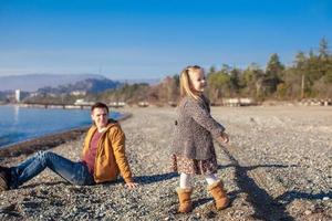 Entzückendes kleines Mädchen mit Vater, das sich am warmen Wintertag am Strand amüsiert foto