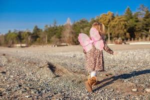 entzückendes kleines Mädchen mit Schmetterlingsflügeln, das an einem sonnigen Wintertag am Strand entlang läuft foto