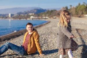 Entzückendes kleines Mädchen mit Vater, das sich am warmen Wintertag am Strand amüsiert foto