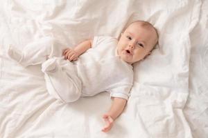 Porträt eines süßen Babys in einem weißen Body auf einem Bett zu Hause mit weißer Bettwäsche. neugeborenes baby zu hause, glückliche mutterschaft. hochwertiges Foto