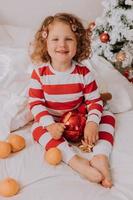 kleines, lockiges, fröhliches mädchen im weihnachtspyjama isst lollipop und mandarinen, die im bett sitzen. Kind genießt Weihnachten. Lebensstil. Platz für Text. hochwertiges Foto