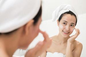 glückliche junge asiatin, die gesichtslotionen aufträgt, während sie ein handtuch trägt und ihr gesicht im badezimmer berührt foto