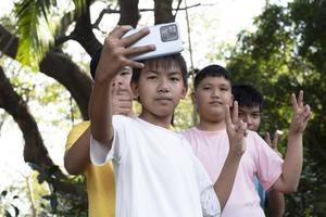 Gruppe junger asiatischer Teenager, die Freizeit im Park verbringen, ihre Finger heben und glücklich zusammen Selfie machen, weicher und selektiver Fokus auf Jungen im weißen T-Shirt, Teenager-Konzept aufziehen. foto