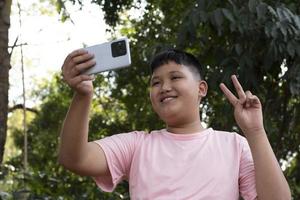 Gruppe junger asiatischer Teenager, die Freizeit im Park verbringen, ihre Finger heben und glücklich zusammen Selfie machen, weicher und selektiver Fokus auf Jungen im weißen T-Shirt, Teenager-Konzept aufziehen. foto