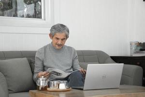 porträt älterer asiatischer mann sitzt morgens in der nähe eines glasfensters, um von zu hause aus zu arbeiten und überprüft sein geschäft auf seinem laptop auf dem tisch ernsthaft, weicher und selektiver fokus. foto