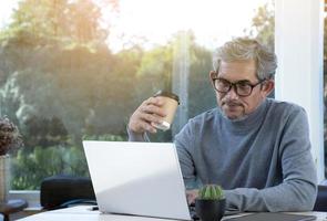 porträt älterer asiatischer mann sitzt morgens in der nähe eines glasfensters, um von zu hause aus zu arbeiten und überprüft sein geschäft auf seinem laptop auf dem tisch ernsthaft, weicher und selektiver fokus. foto