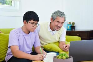 Der asiatische Senior-Opa und sein Sohn verbringen Zeit zusammen zu Hause, um Kopfhörer mit seinem Laptop zu verbinden, um Online-Spiele anzusehen und zu spielen, weicher und selektiver Fokus, Teenager-Konzept aufziehen. foto