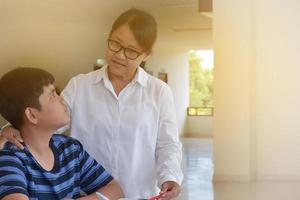 junger asiatischer junge macht seine aufgabe und hört sich die ratschläge seiner älteren lehrerin zum schulprojekt an, erwachsener hilft kindern beim konzept des schulprojekts. foto