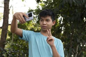 Gruppe junger asiatischer Teenager, die Freizeit im Park verbringen, ihre Finger heben und glücklich zusammen Selfie machen, weicher und selektiver Fokus auf Jungen im weißen T-Shirt, Teenager-Konzept aufziehen. foto
