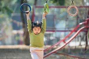 süßes asiatisches mädchenlächeln spielen klettern die bar auf schul- oder kindergartenhof oder spielplatz. gesunde aktivität kinder. kleines Mädchen, das draußen auf dem Spielplatz klettert. Kind spielt auf Spielplatz im Freien. foto