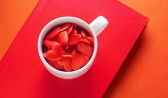 liebesgetränk, weiße tasse voller herzen auf rotem buch und orangefarbenem hintergrund, valentinstagkonzept mit lebendigen farben foto