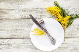 festlicher tischgedeckteller mit gabel und messer und mimosenblumendekoration auf weißem hölzernem hintergrund mit kopienraum foto