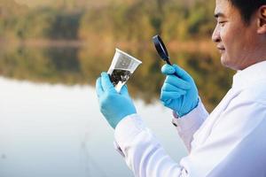 nahaufnahme asiatischer ökologe hält lupe, um wasser in transparentem glas aus dem see zu inspizieren. Konzept, Erkundung, Analyse der Wasserqualität und Lebewesen aus natürlichen Quellen. Ökologische Feldforschung foto