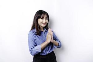 lächelnde junge asiatische frau, die blaues hemd trägt und traditionellen gruß gestikuliert, der über weißem hintergrund lokalisiert wird foto
