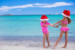 kleine süße mädchen in weihnachtsmützen, die spaß am exotischen strand haben foto