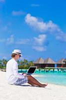 Junger Mann, der am tropischen Strand in der Nähe der Wasservilla am Laptop arbeitet foto