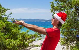 junger Mann mit Weihnachtsmütze am tropischen Strand in der Nähe der Tannen foto