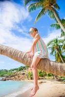 entzückendes kleines mädchen, das während der sommerferien auf der palme am weißen strand sitzt foto