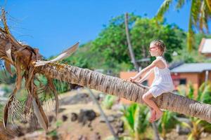 entzückendes kleines mädchen, das während der sommerferien auf der palme am weißen strand sitzt foto