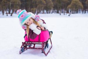 Entzückendes kleines glückliches Mädchen, das im verschneiten Wintertag Rodeln fährt foto