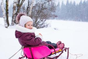 Entzückendes kleines glückliches Mädchen, das im verschneiten Wintertag Rodeln fährt foto