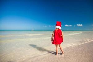 Rückansicht des kleinen süßen Mädchens mit rotem Hut Weihnachtsmann am Strand foto