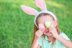 Entzückendes kleines Mädchen, das am Frühlingstag Hasenohren mit Ostereiern trägt foto