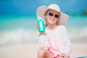 Kleines Mädchen mit Hut und einer Flasche Sonnencreme, die auf der Sonnenliege am tropischen Strand sitzt foto