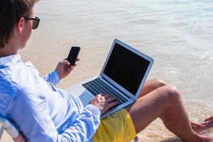 junger Geschäftsmann, der Laptop und Telefon am tropischen Strand benutzt foto