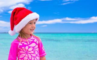 kleines entzückendes Mädchen mit Weihnachtsmütze am tropischen Strand foto