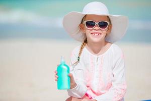 Kleines Mädchen mit Hut und einer Flasche Sonnencreme, die auf der Sonnenliege am tropischen Strand sitzt foto