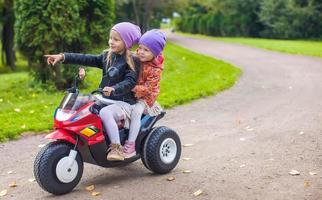 kleine entzückende schwestern, die auf spielzeugmotorrad im grünen park sitzen foto