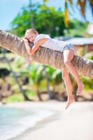 Entzückendes kleines Mädchen am tropischen Strand, das während der Sommerferien auf Palmen sitzt foto
