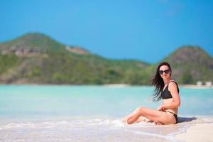 Frau, die lachend am Strand sitzt und die Sommerferien genießt und in die Kamera schaut. schönes Modell im Bikini, das sich hinsetzt. foto