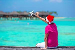 junger Mann mit Weihnachtsmütze am weißen Strand mit Miniatur des Flugzeugs foto