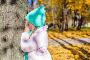 Kleines Mädchen, das Verstecken spielt und in der Nähe des Baums im Herbstpark sucht foto